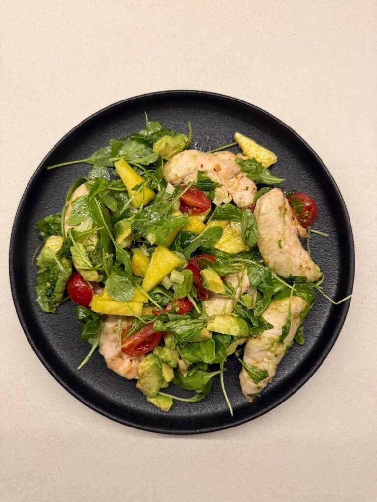 Ensalada de pollo con aguacate, mango y rúcula en un plato negro.